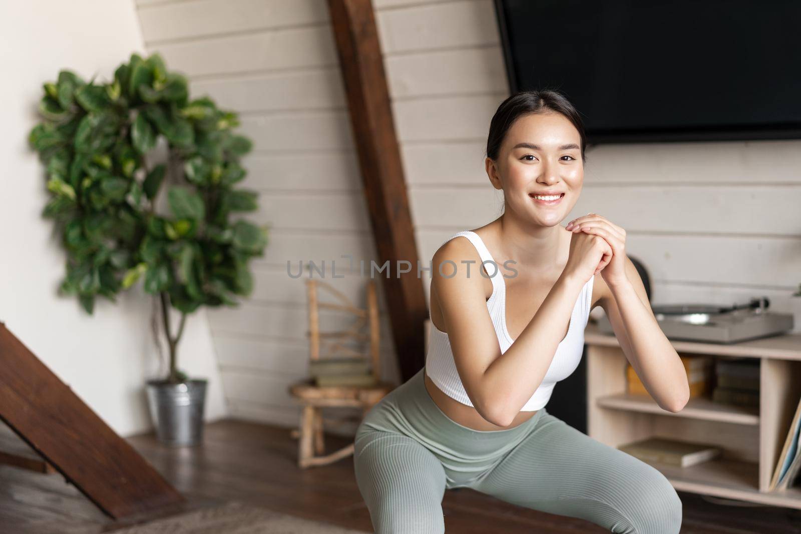 Young asian fitness woman with healthy fit body, doing squats, morning workout, wearing activewear, standing at home in living room and having training session by Benzoix