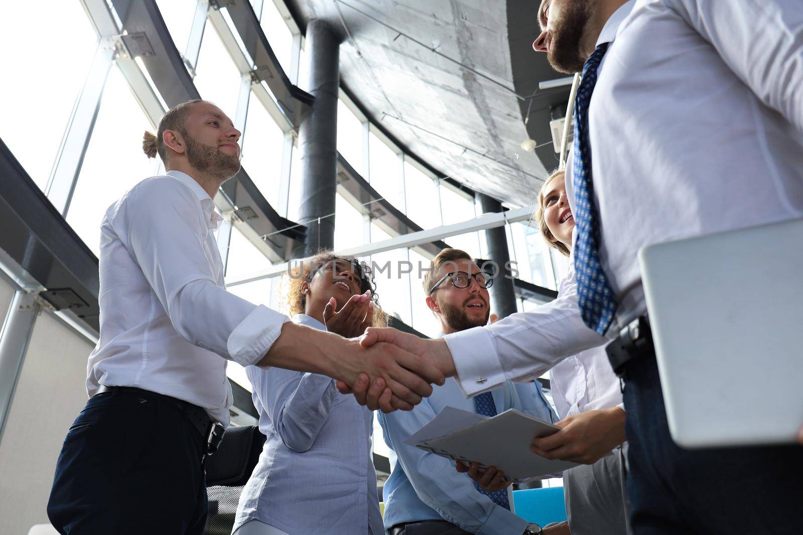 Two smiling businessmen are shaking hands while being in office together with their colleagues.