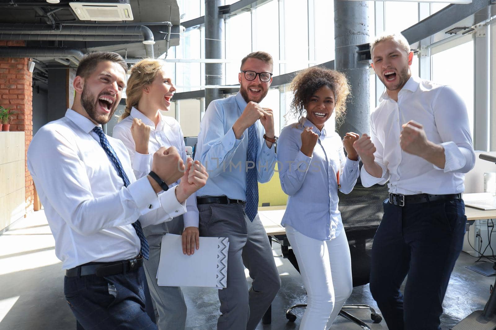 Five happy modern business people are keeping arms raised and expressing joyful while standing in large office.