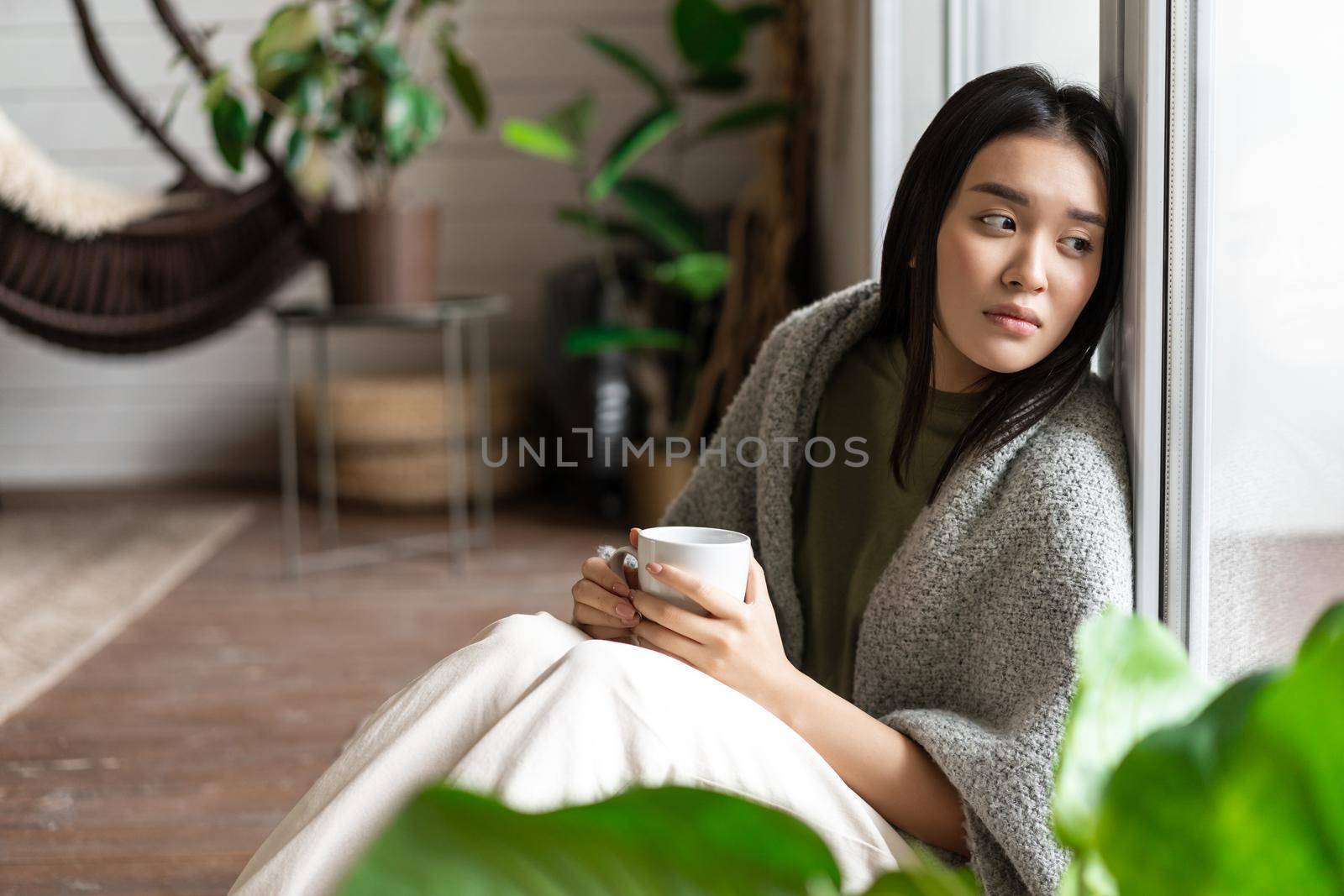 Sad asian woman looking outside window, sitting on floor with miserable upset face, drinking coffee at home.