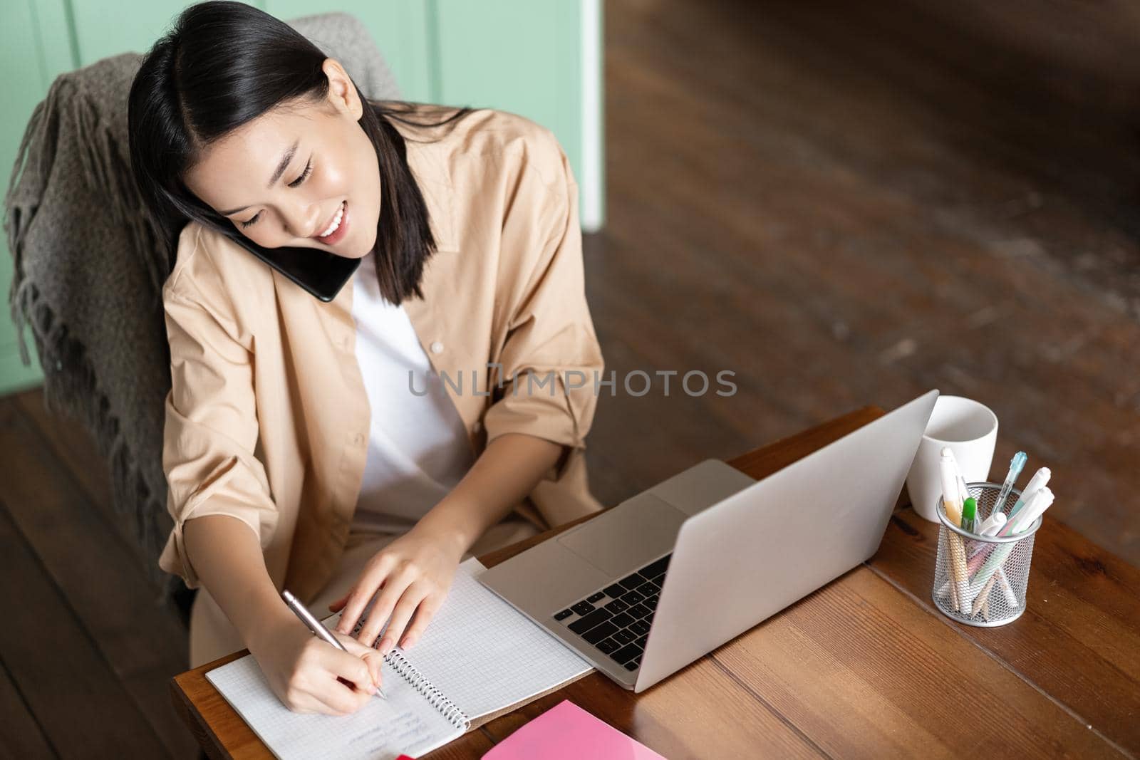 Upper angle shot of asian woman working with laptop, answer phone call and writing down in notebook. Girl takes notes while talking on smartphone, sitting near computer.