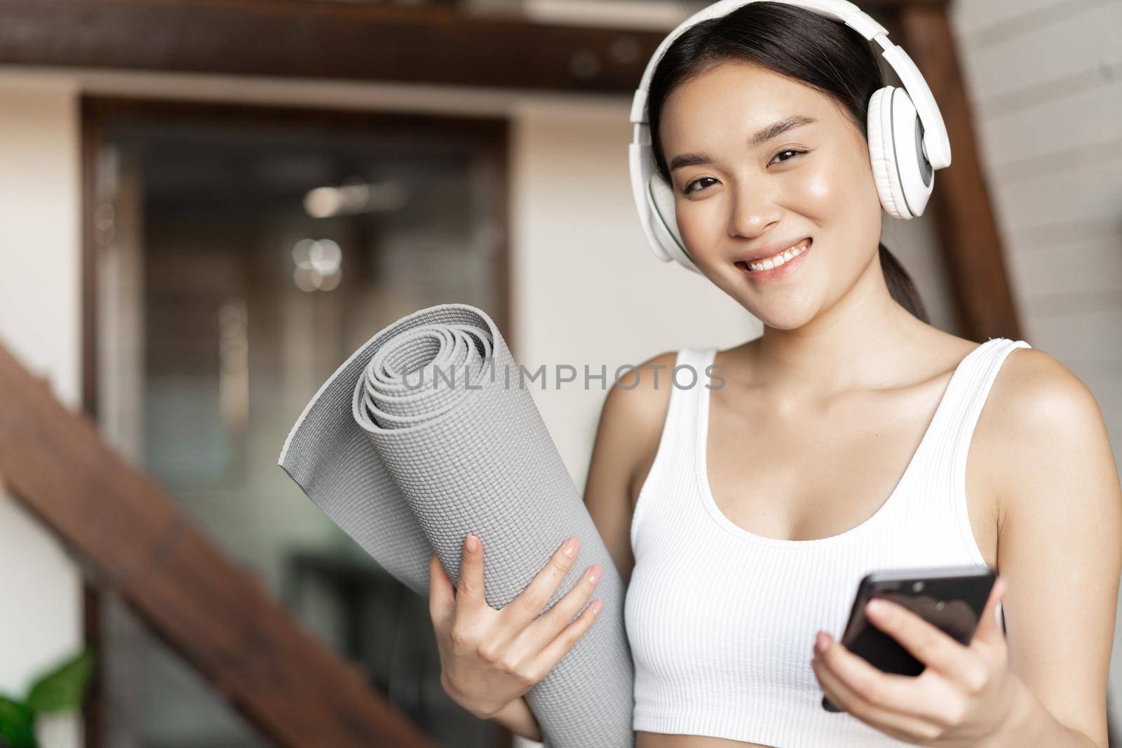 Smiling asian girl in headphones, listening music at home and workout with floor mat, holding mobile phone, fitness exercises in living room.