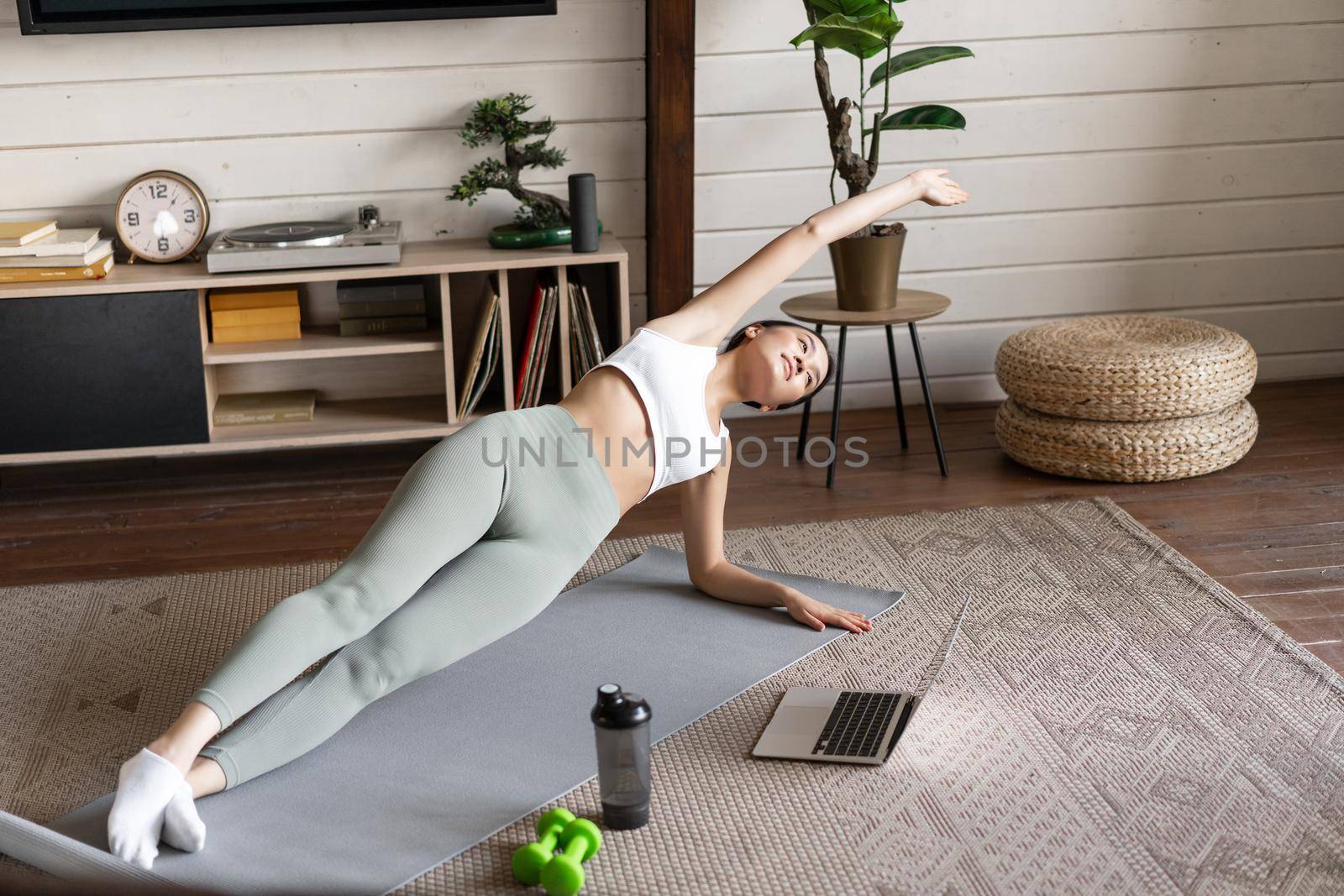 Young asian woman doing workout at home on floor mat, following sport instructor on laptop video, stretching arm and doing plank.