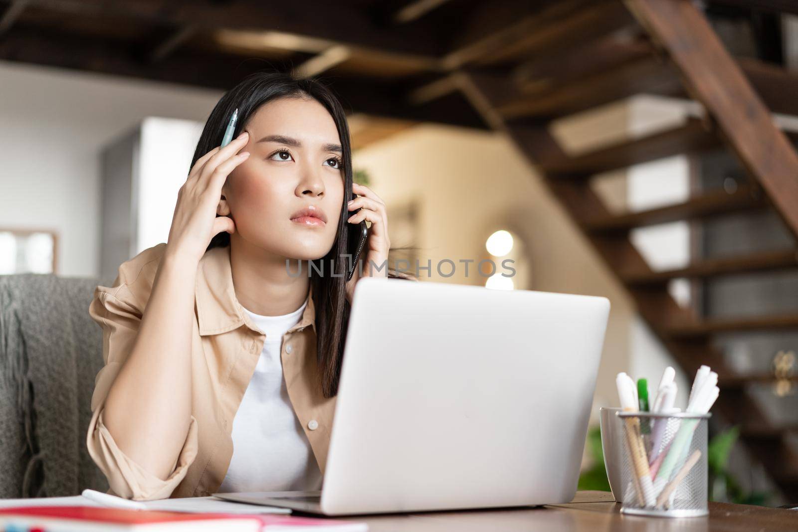 Asian woman working from home, answer phone call and listen with serious face client, sitting near laptop.