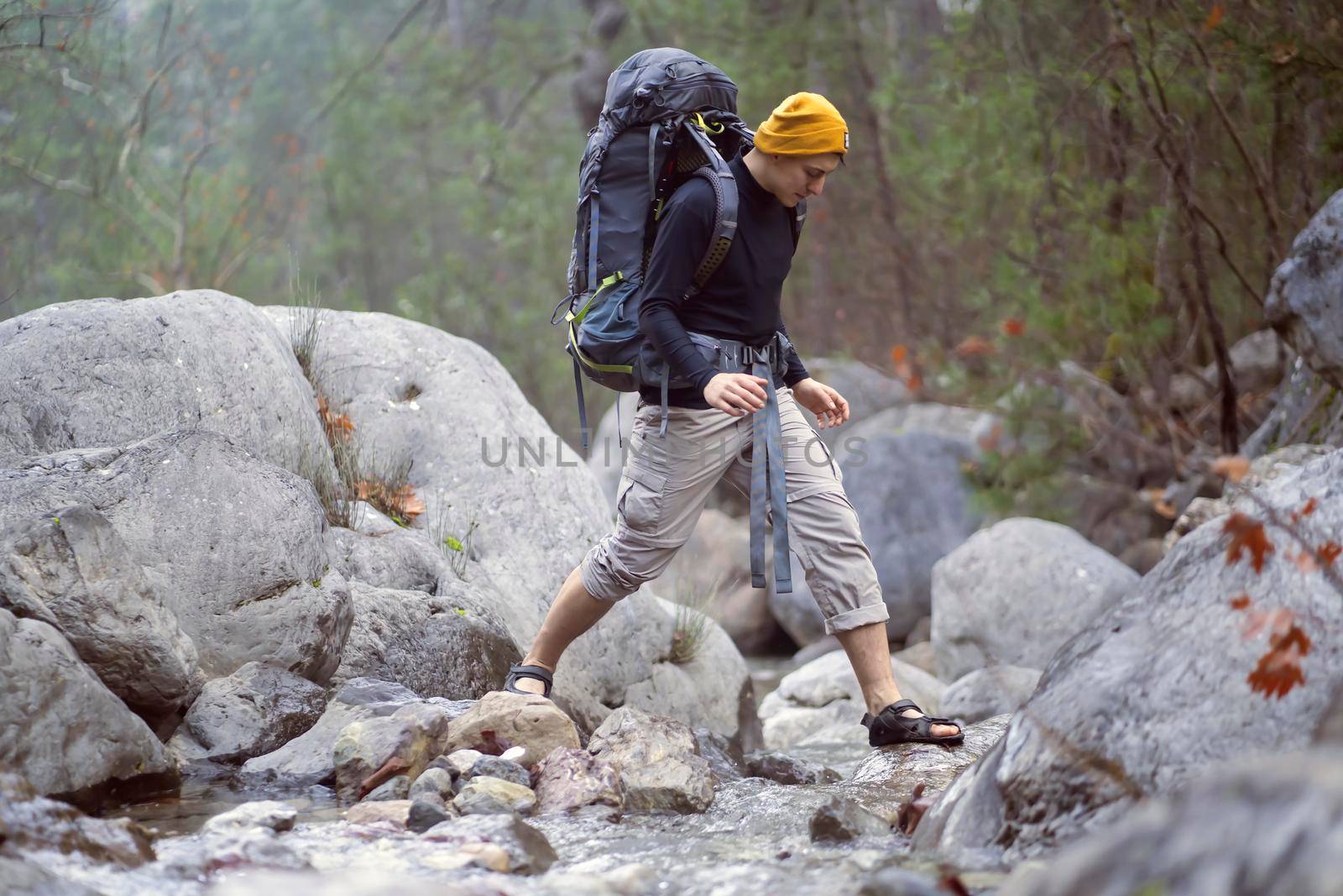 Young man with backpack and in hiking clothes carefully crosses the river in the mountains. Stylish traveler doing trekking along the trail in the national park, it is surrounded by rocks and forests.