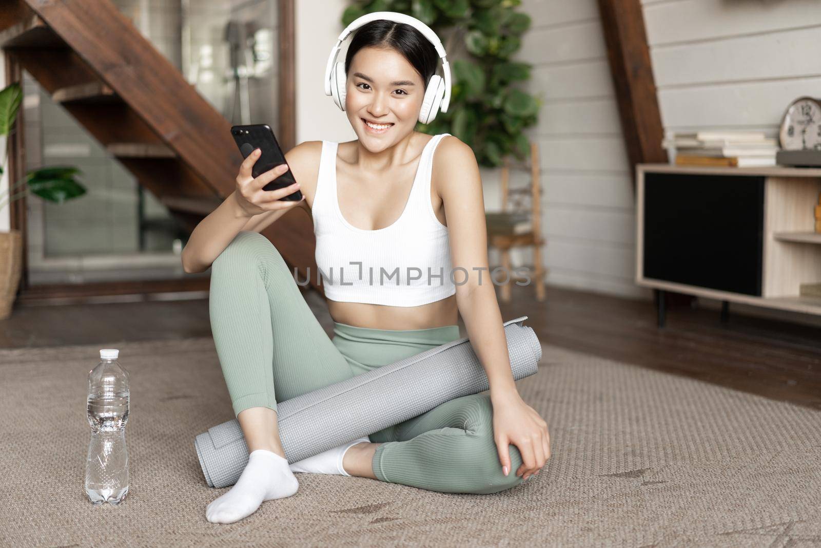 Smiling asian girl in headphones wearing sport clothing, listening music, holding mobile phone and workout at home alone, sitting in living room by Benzoix