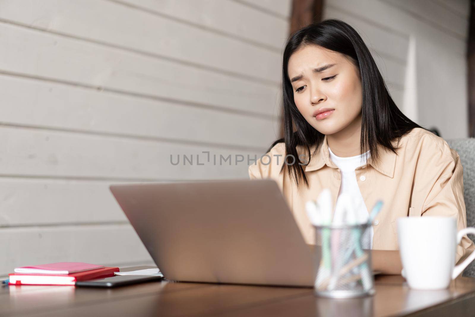 Sad and tired asian girl looking disappointed at laptop screen, bored of studying, watching something boring on computer.