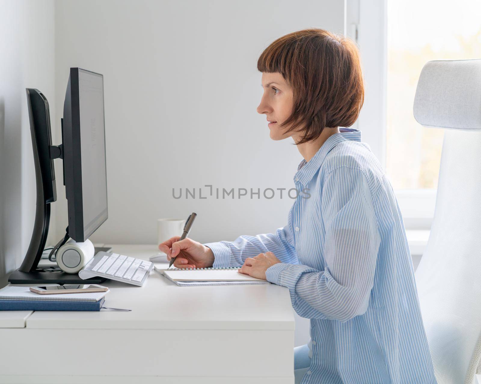 Mature woman looking at computer screen and writing information in notebook. Online course, Education. Side view of middle aged female working remotely from home