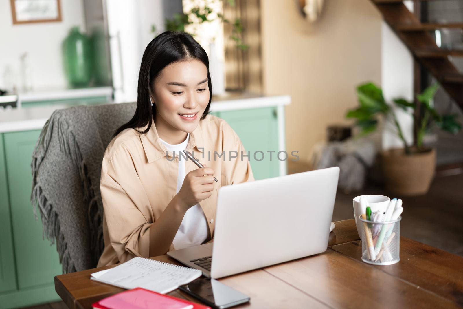 Smiling asian girl studying at home on laptop, listening webinar and taking notes, writing down homework with computer.