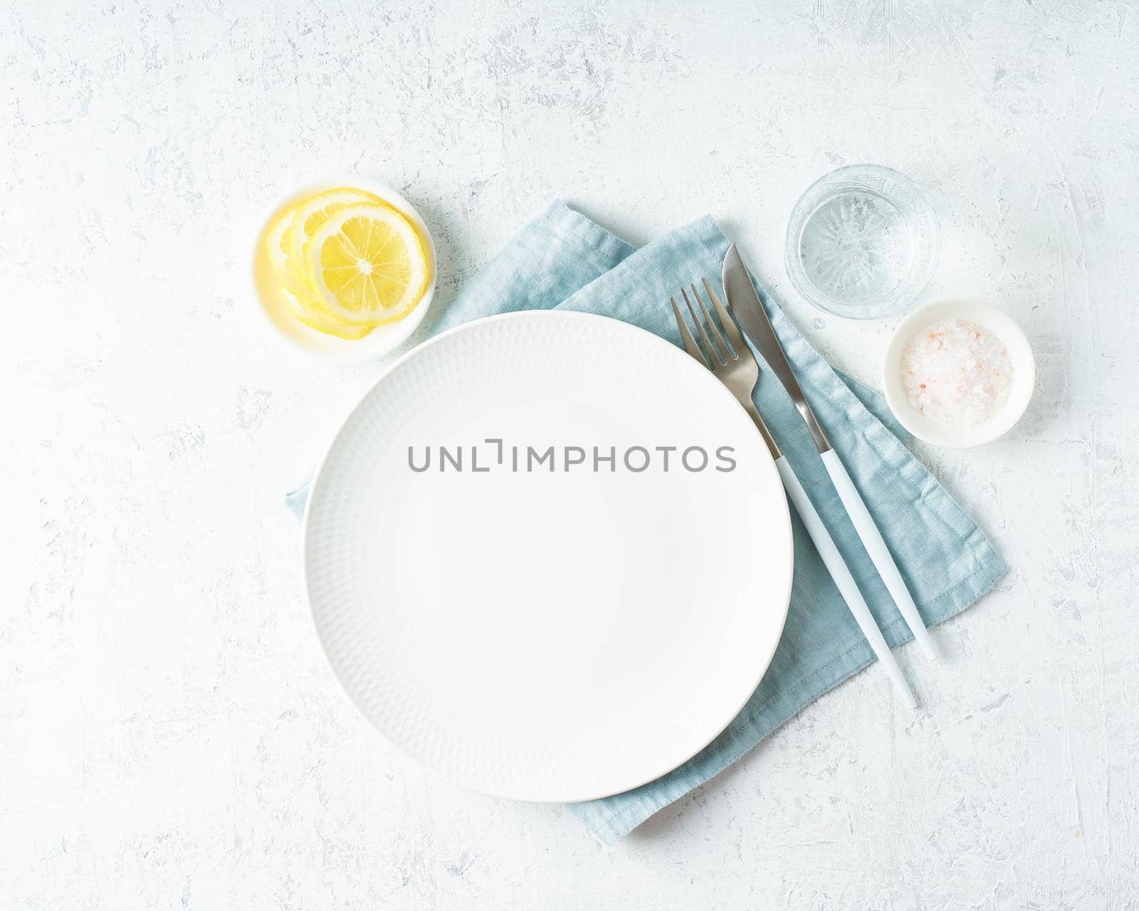 Clean empty white plate, glass of water, fork and knife on white stone table, copy space, mock up, top view. Concept for menu with utensil