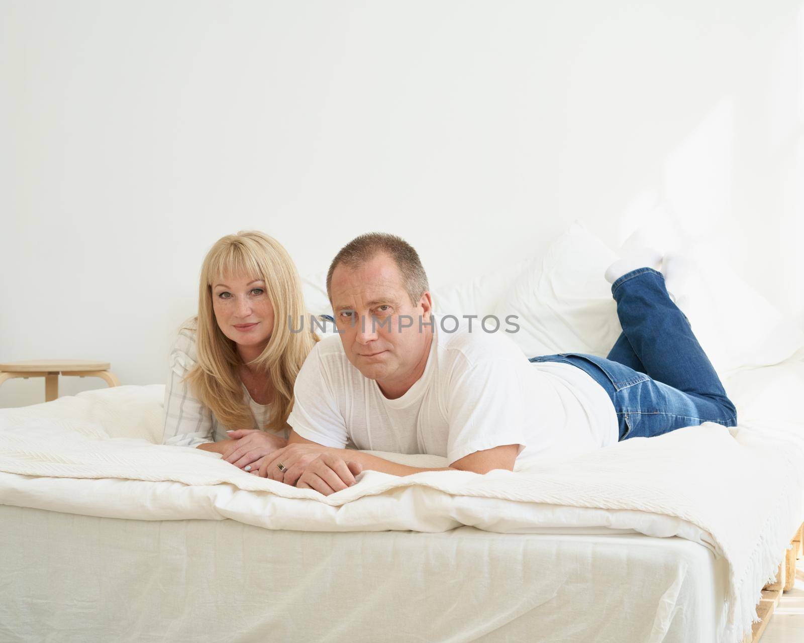 Portrait of mature couple in home interior on sofa. Handsome man and attractive middle age woman enjoying spending time together while lying in bed. People looking at camera