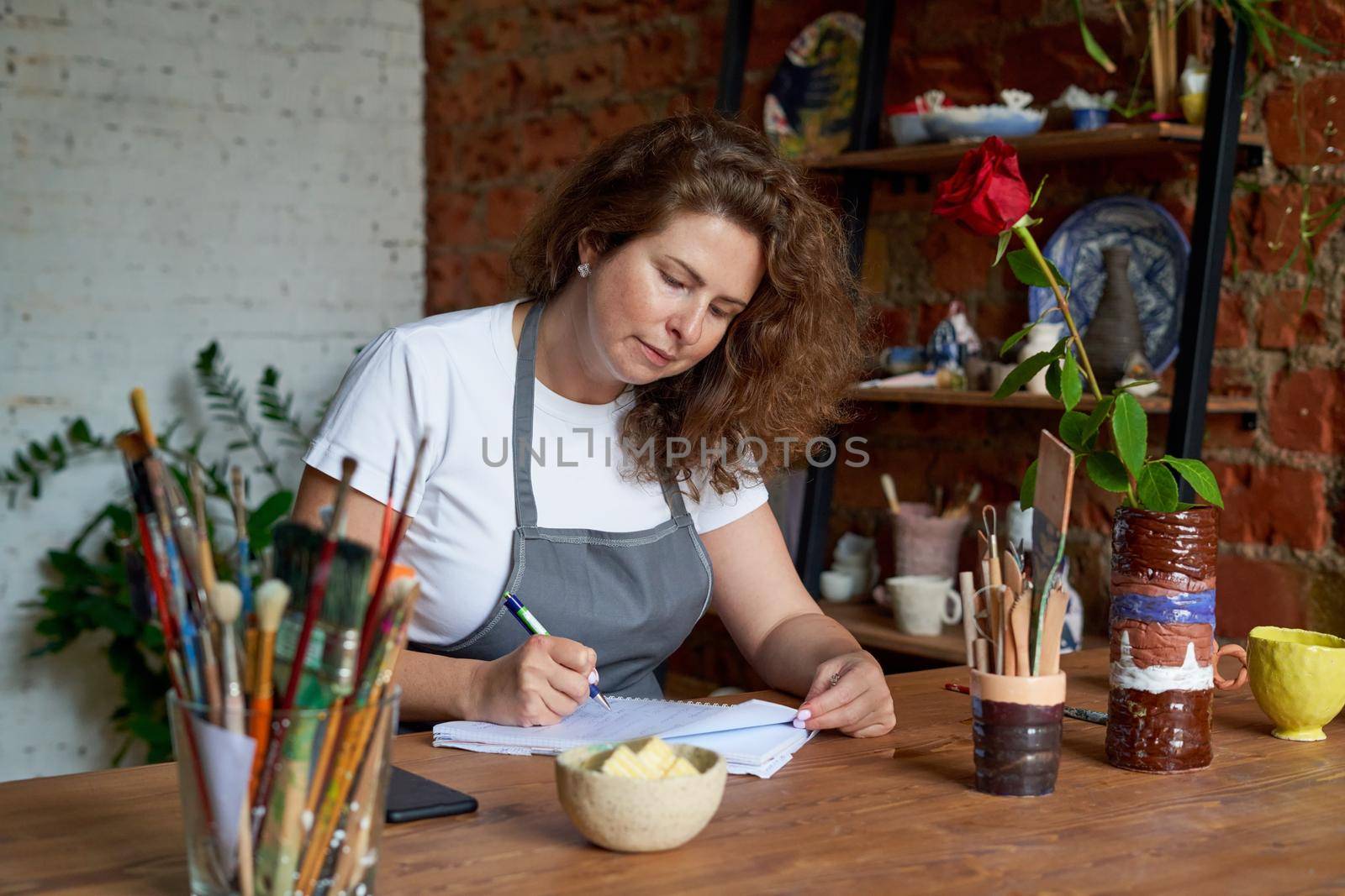 Artisan woman writing n notebook, planning day in diary. Beautiful business female entrepreneur with long curly hair sits at workshop in chair and thinking about working on projects