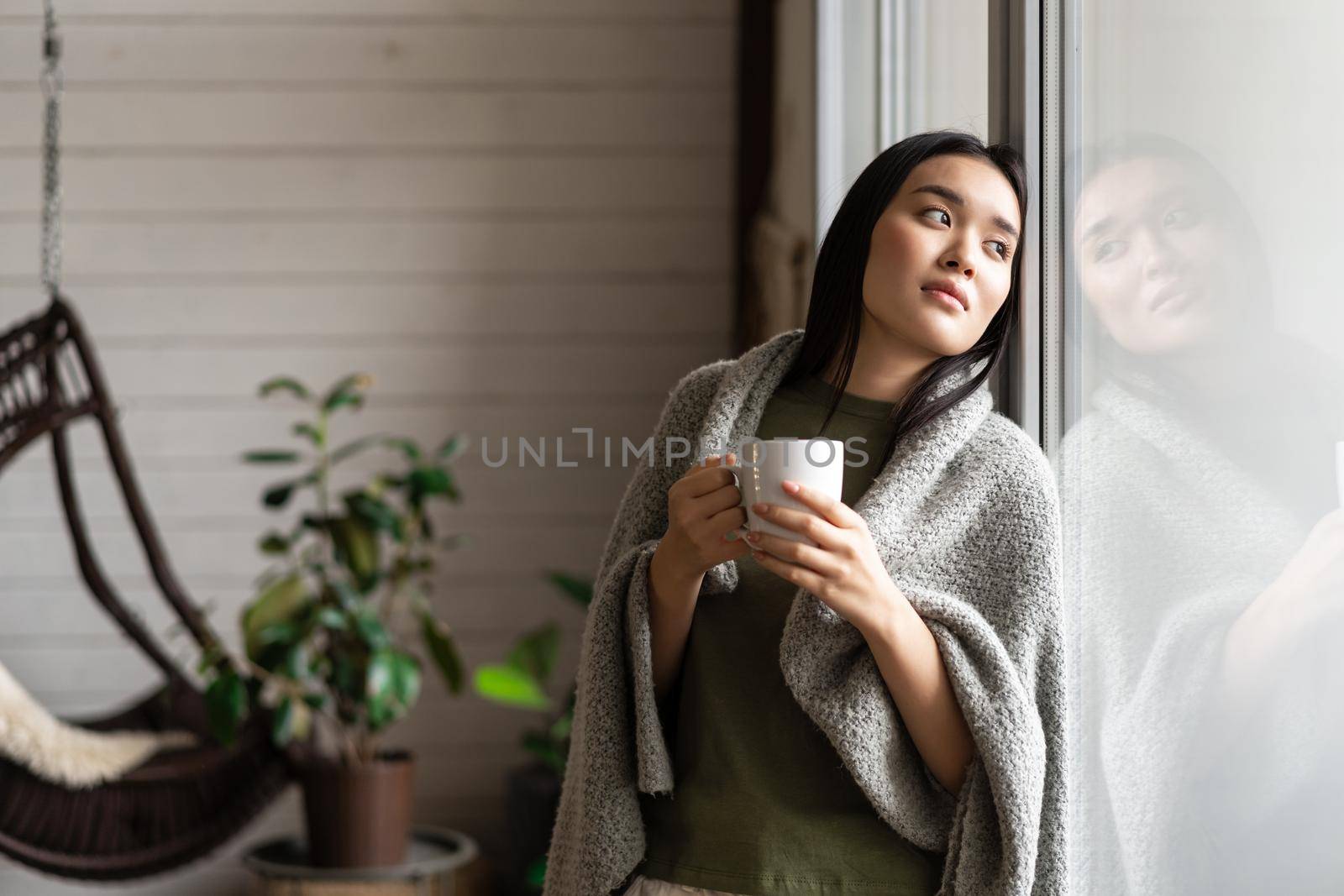 Romantic asian woman wrapped in blanket, leaning on window and looking outside, drinking hot coffee. Lifestyle concept