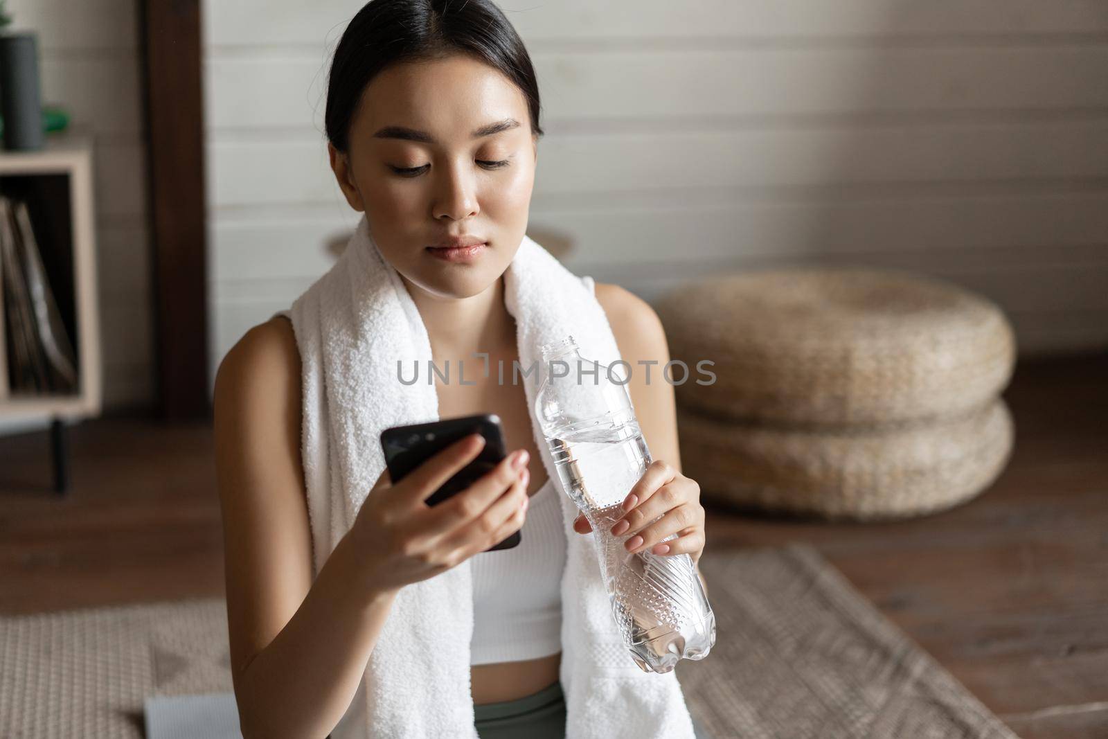 Image of young asian fitness girl with fit body, towel wrapped around neck, looking at mobile phone app, checking application during workout.