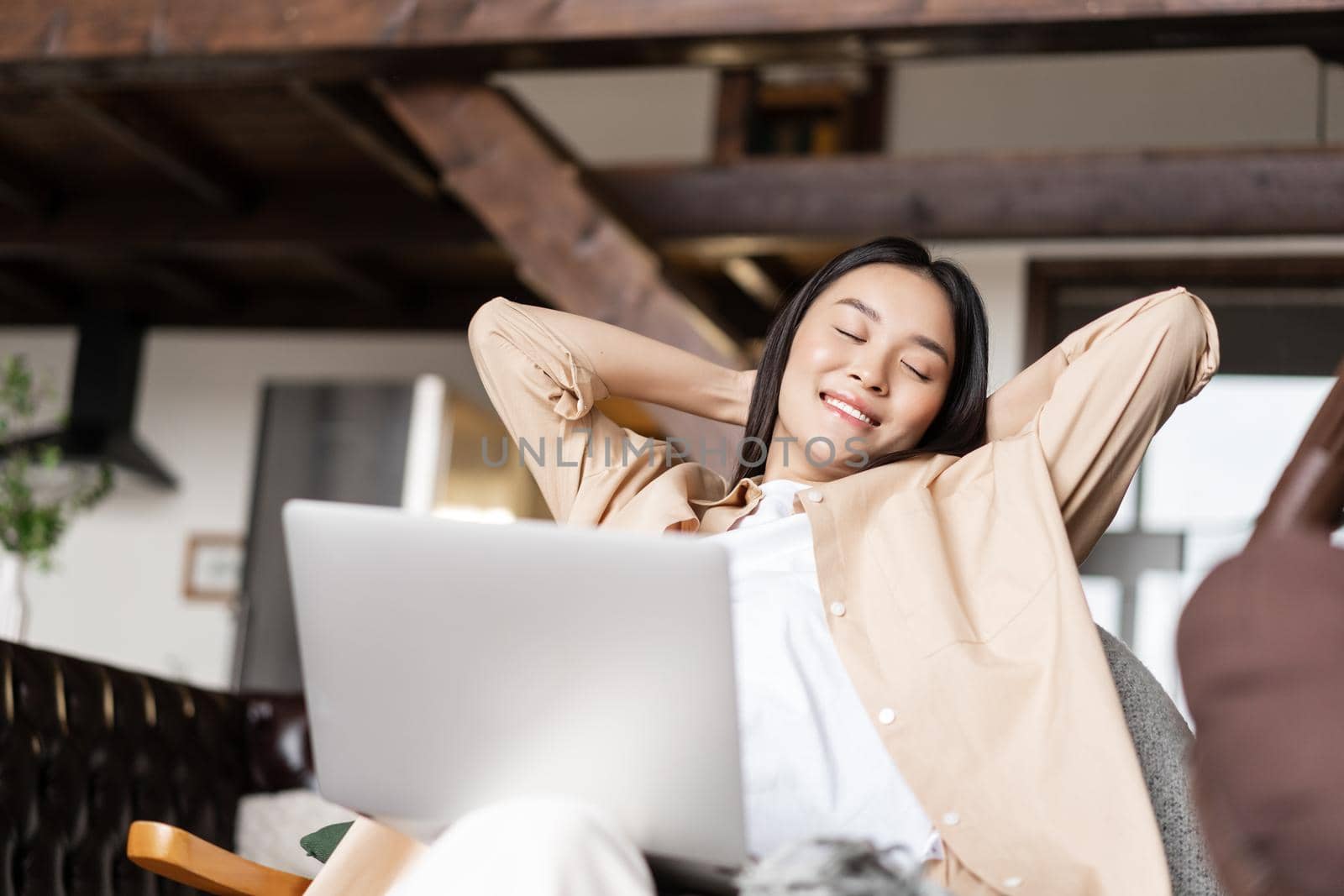 Smiling korean woman resting with hands behind her head, using laptop and relaxing at home by Benzoix