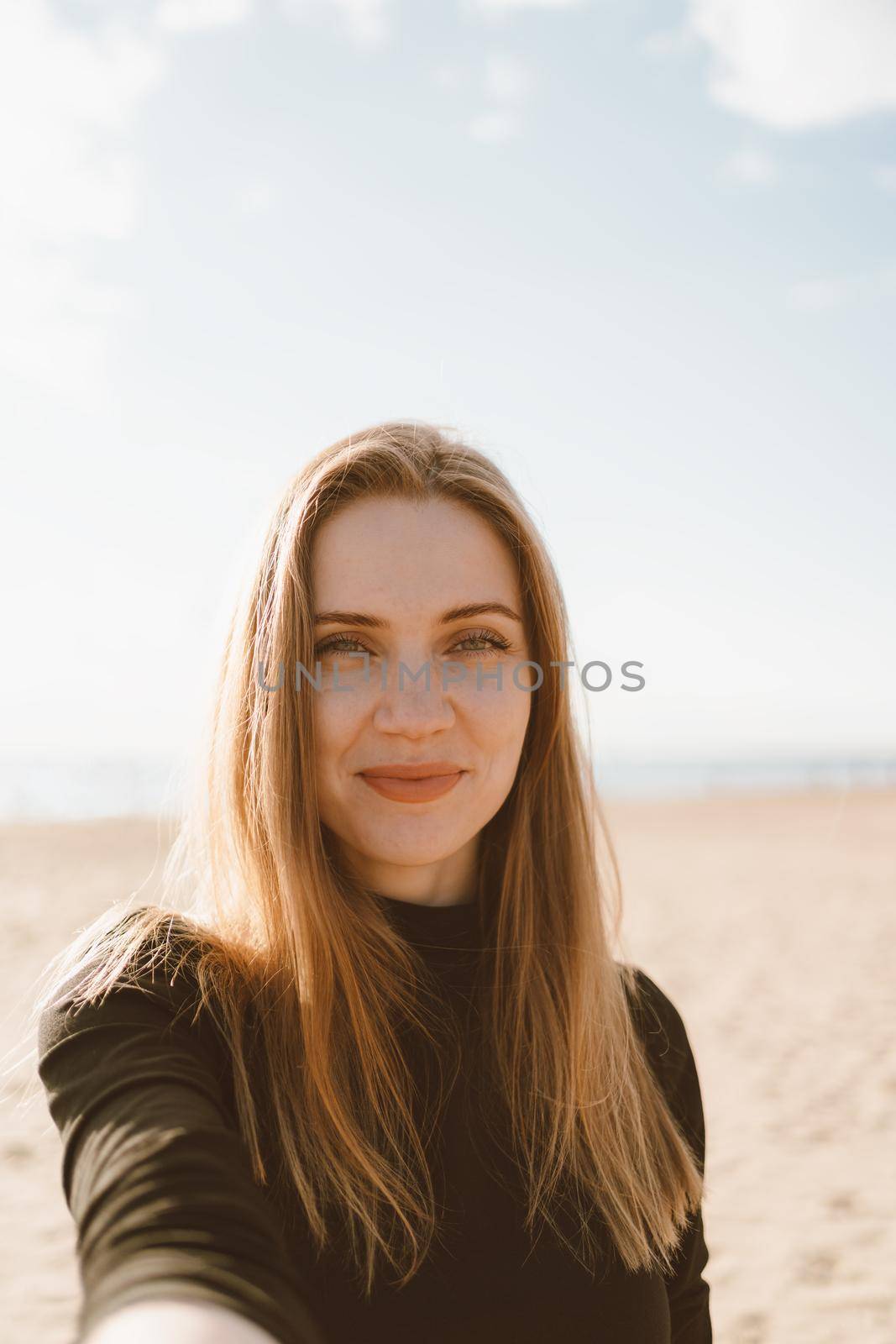 Portrait of pretty female with long hair, blonde takes selfie on mobile phone on sandy beach by NataBene