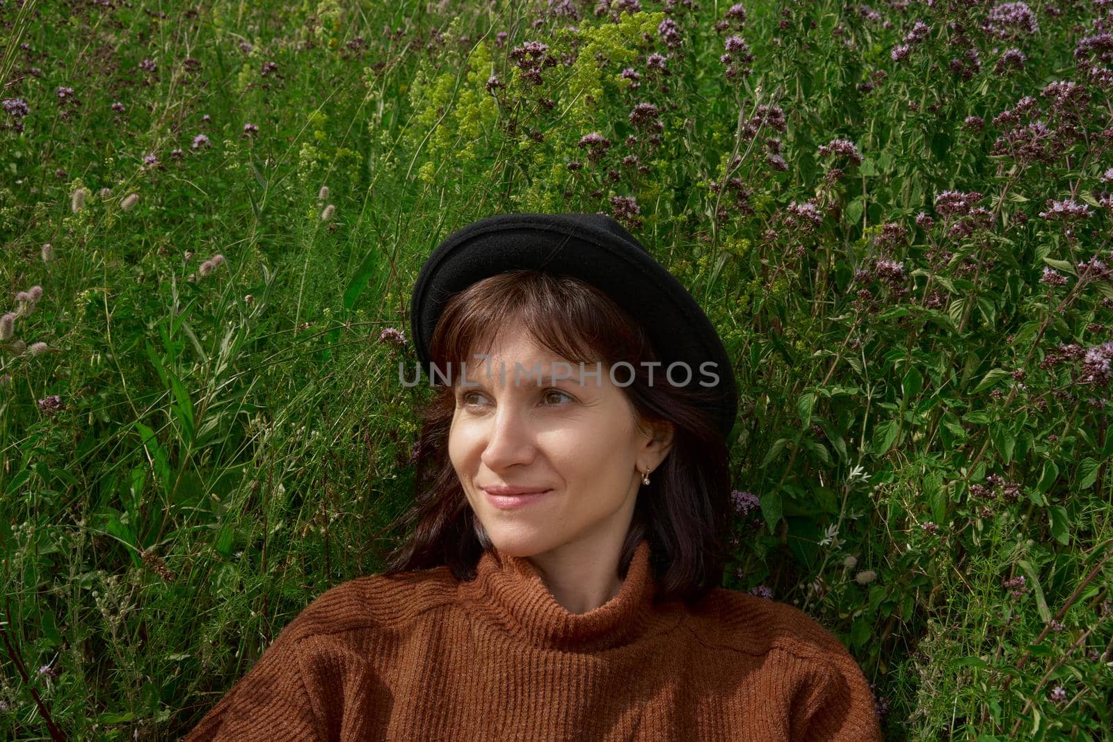 Horizontal portrait of brunette smiling and looking away young adult lady wearing hat and sweater sitting in wildflower field Karelia