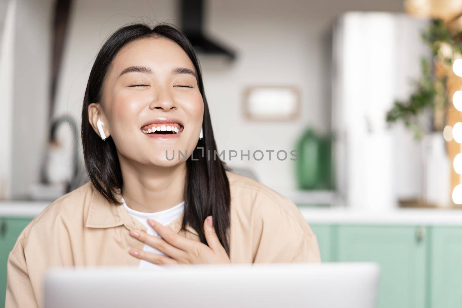 Happy asian girl laughing, watching video or having a call via laptop application, working remotely from home during pandemic.