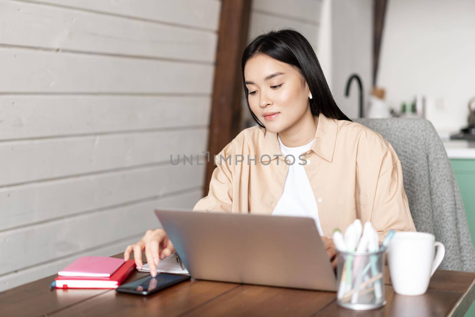 Young asian girl checking smartphone app while working on laptop, using devices while doing homework or project on computer, workplace at home concept.