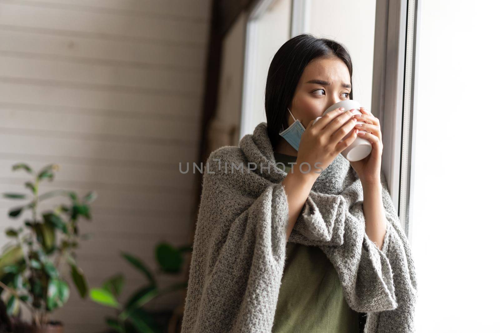 Sick asian girl drinking hot tea, staying on quarantine at home, looking outside window, feeling ill and sad.