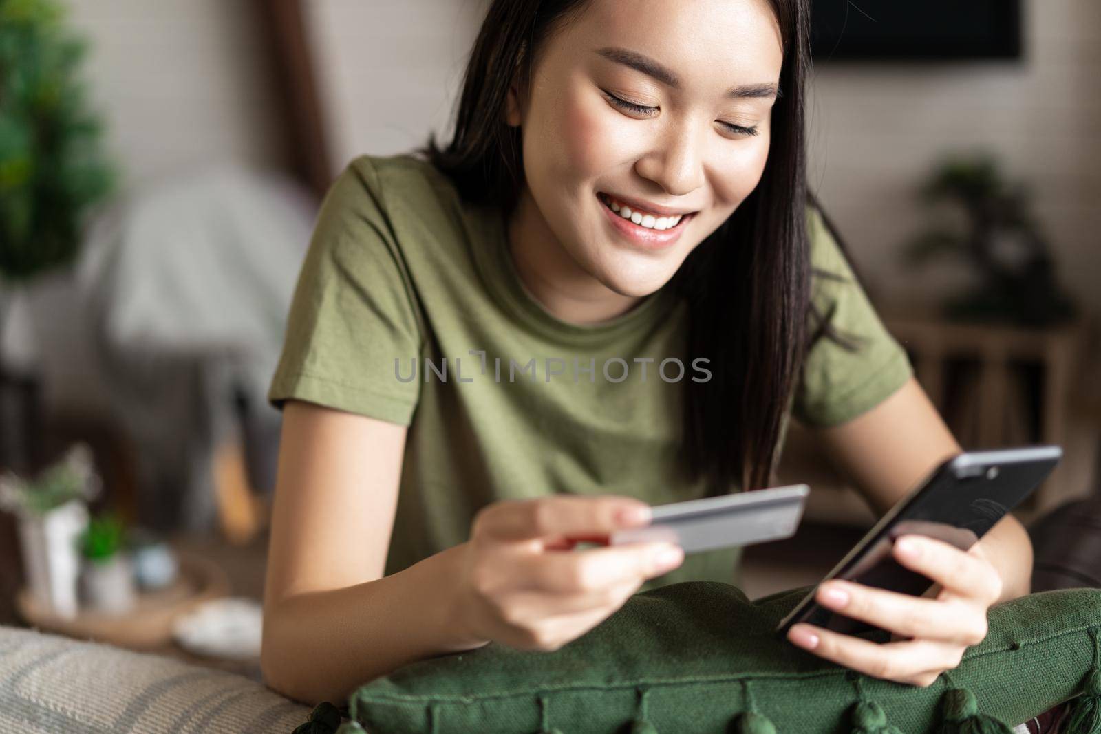 Online shopping. Young asian woman sitting at home and making purchase on mobile phone app, holding credit card.
