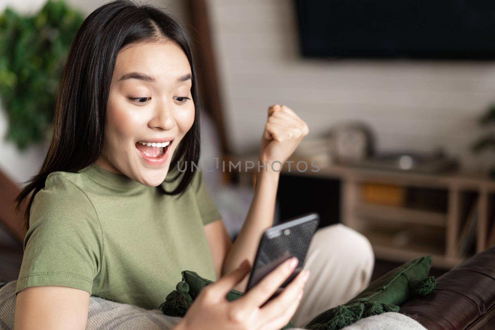 Smiling asian woman looking satisfied, making fist pump and reading mobile phone, sitting at home and triumphing.