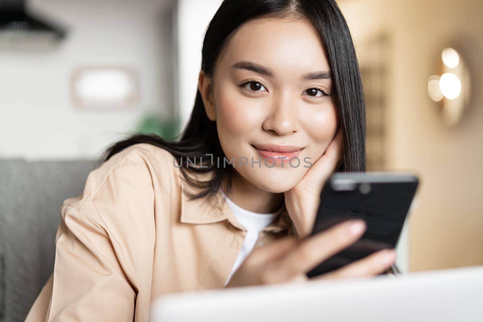 Smiling asian woman working from home on laptop, looking at smartphone.