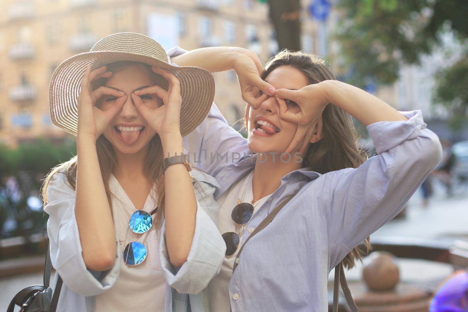 Two young smiling hipster women in summer clothes posing on street.Female showing positive face emotions