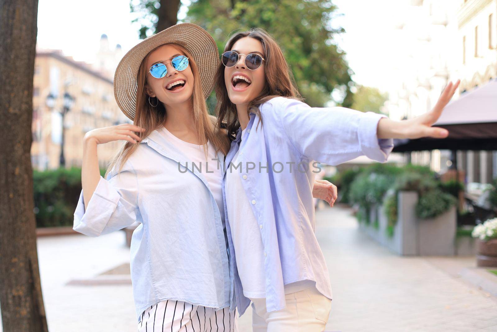 Two young smiling hipster women in summer clothes posing on street.Female showing positive face emotions