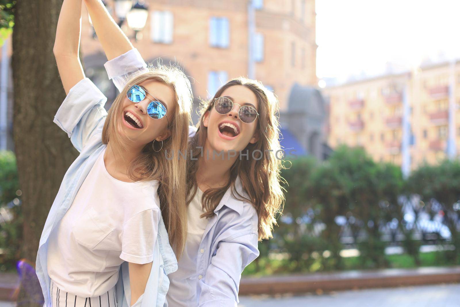 Two beautiful smiling girls in trendy summer clothes posing on street background. Models are having fun and hugging