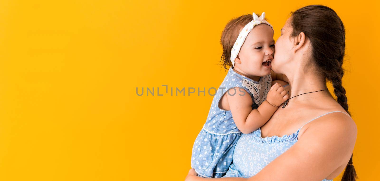 Motherhood, hot summer - Banner young pregnant happy smiling mother woman in blue dress holding little baby daughter toddler sibling in arms looking into eyes kisses on yellow background coppy space.