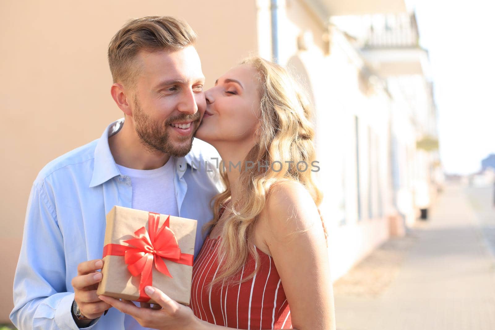 Astonished excited couple in summer clothes smiling and holding present box together while standing on city street.