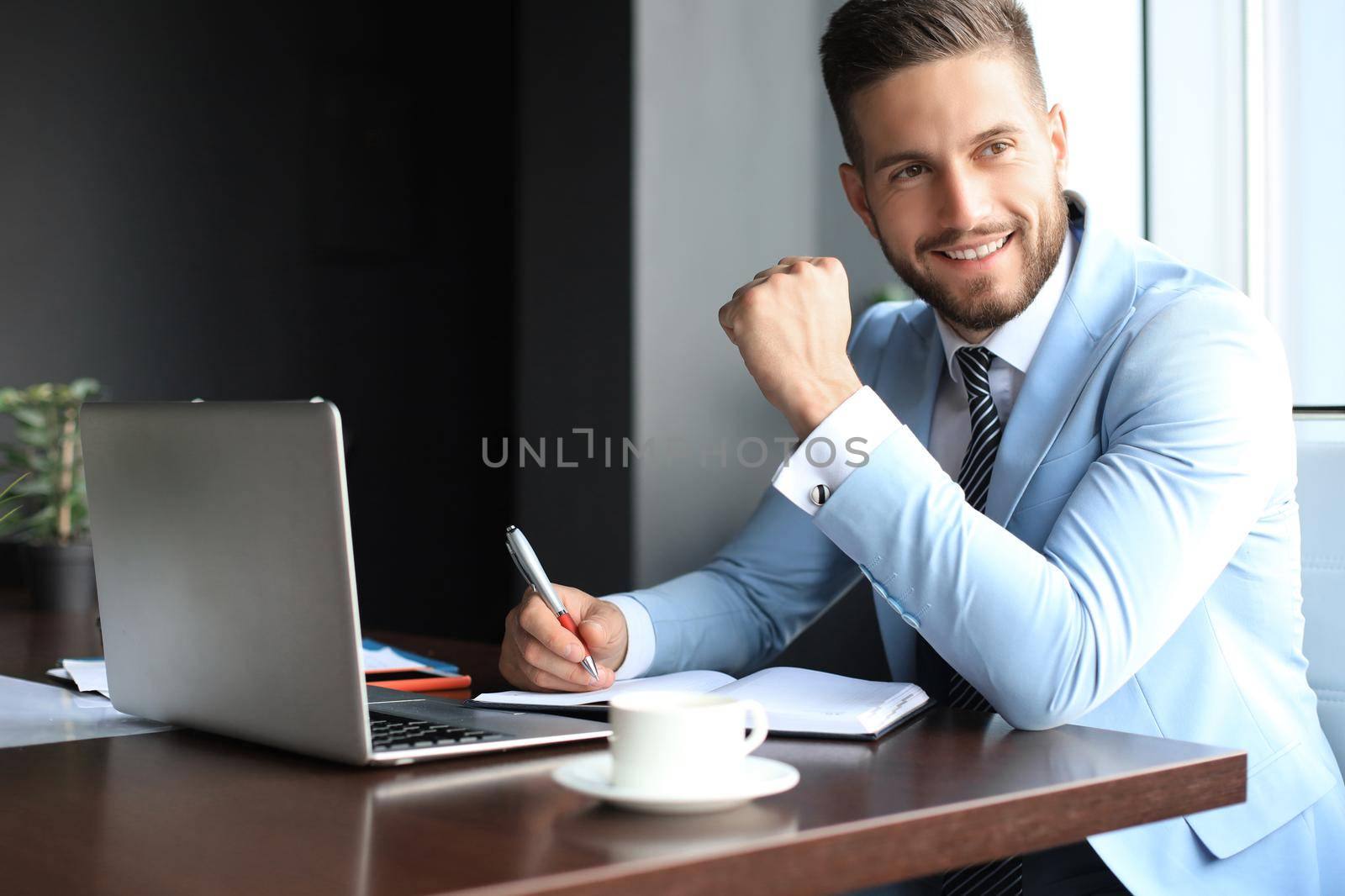 Portrait of happy businessman sitting at office desk, looking at camera, smiling.