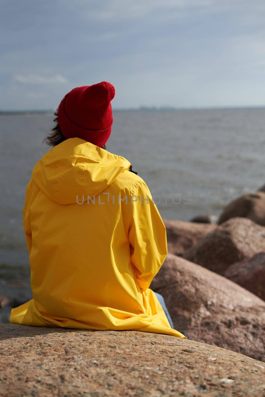 Back view of woman waiting and sitting in bright yellow cloak and red hat on stone near shore of North sea on cold autumn day. Mood Scandinavian weather, deserted beach, coast.