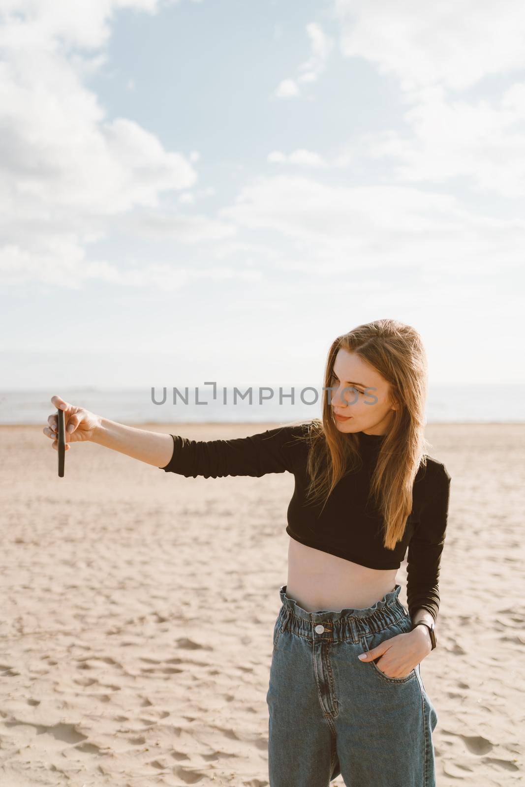 Pretty female with long hair, blonde takes photo on mobile phone on sandy beach in summer or autumn. Beautiful woman in jeans and black top