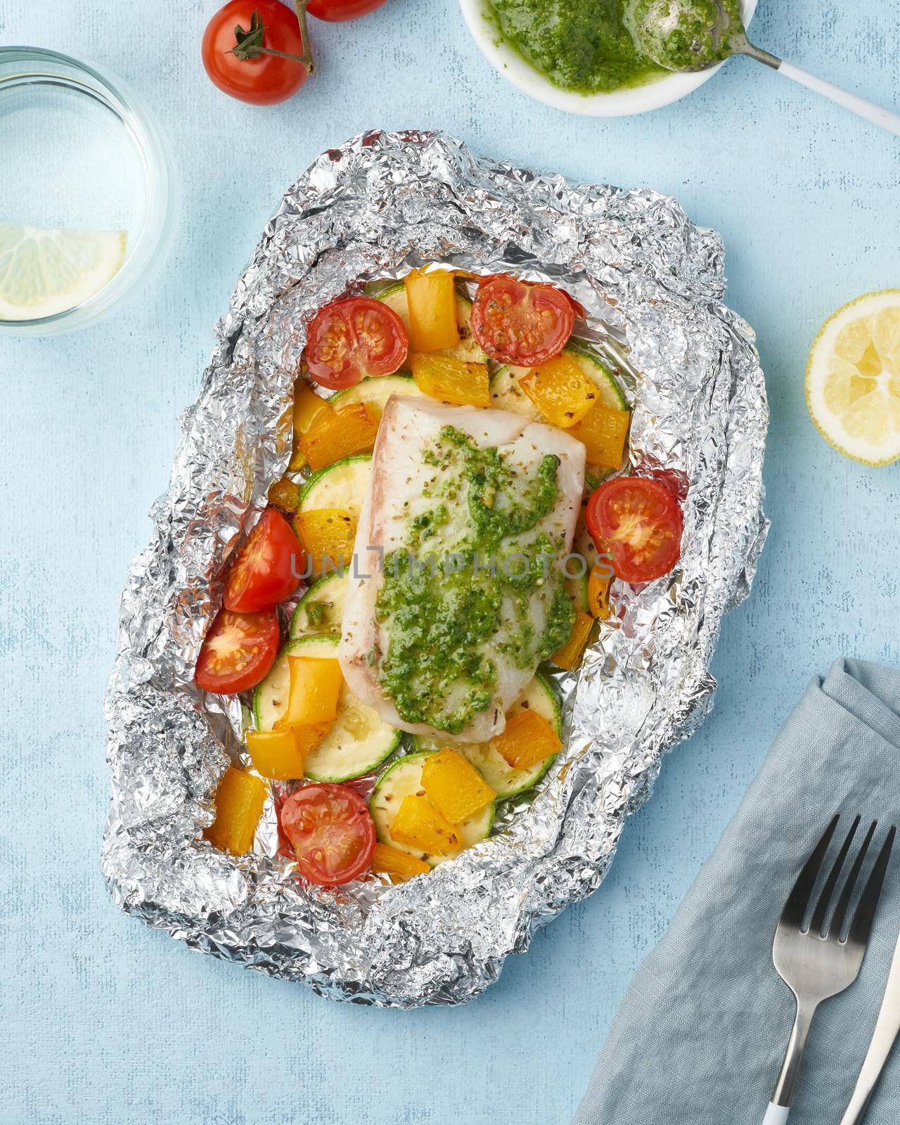 Foil pack dinner with white fish. Oven baked fillet of cod, pike perch with vegetables and pesto sauce. Healthy diet food, keto diet, Mediterranean cuisine. Blue background. Top view, vertical