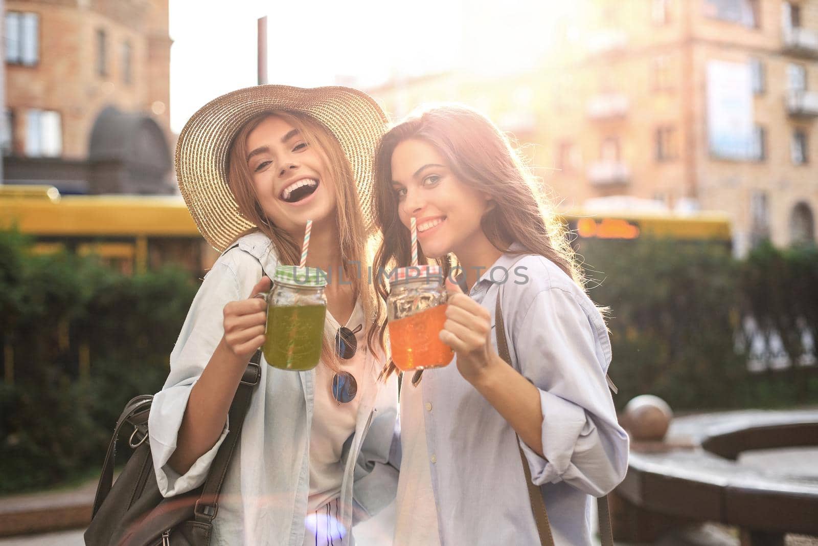 Two young beautiful smiling woman in summer clothes are having fun in city and drinking fresh cocktail