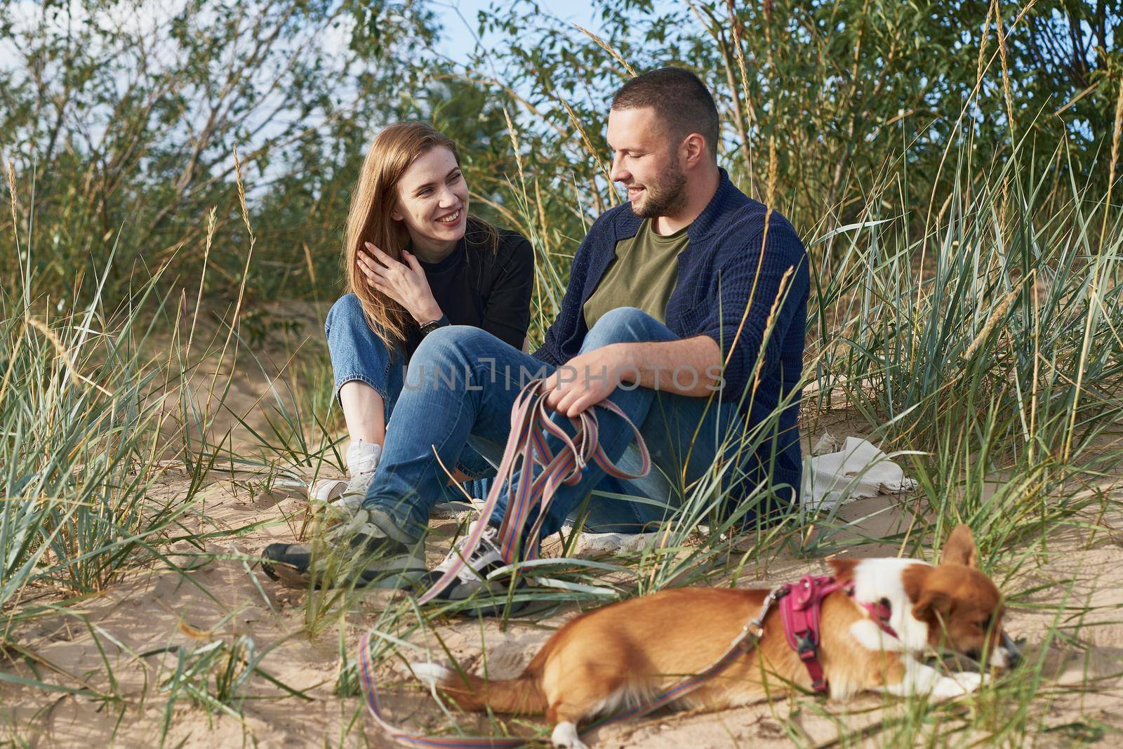 Young happy couple with corgi dog siting at sand. Handsome man and beautiful woman in casual clothes enjoying sunny afternoon on beach