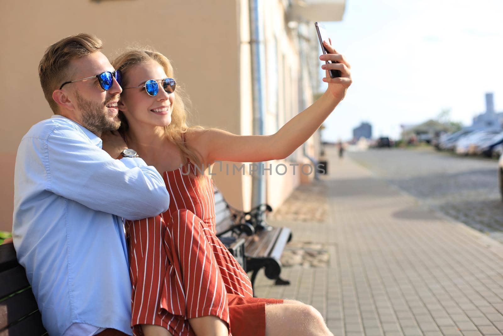 Romantic young couple in summer clothes smiling and taking selfie while sitting on bench in city street.