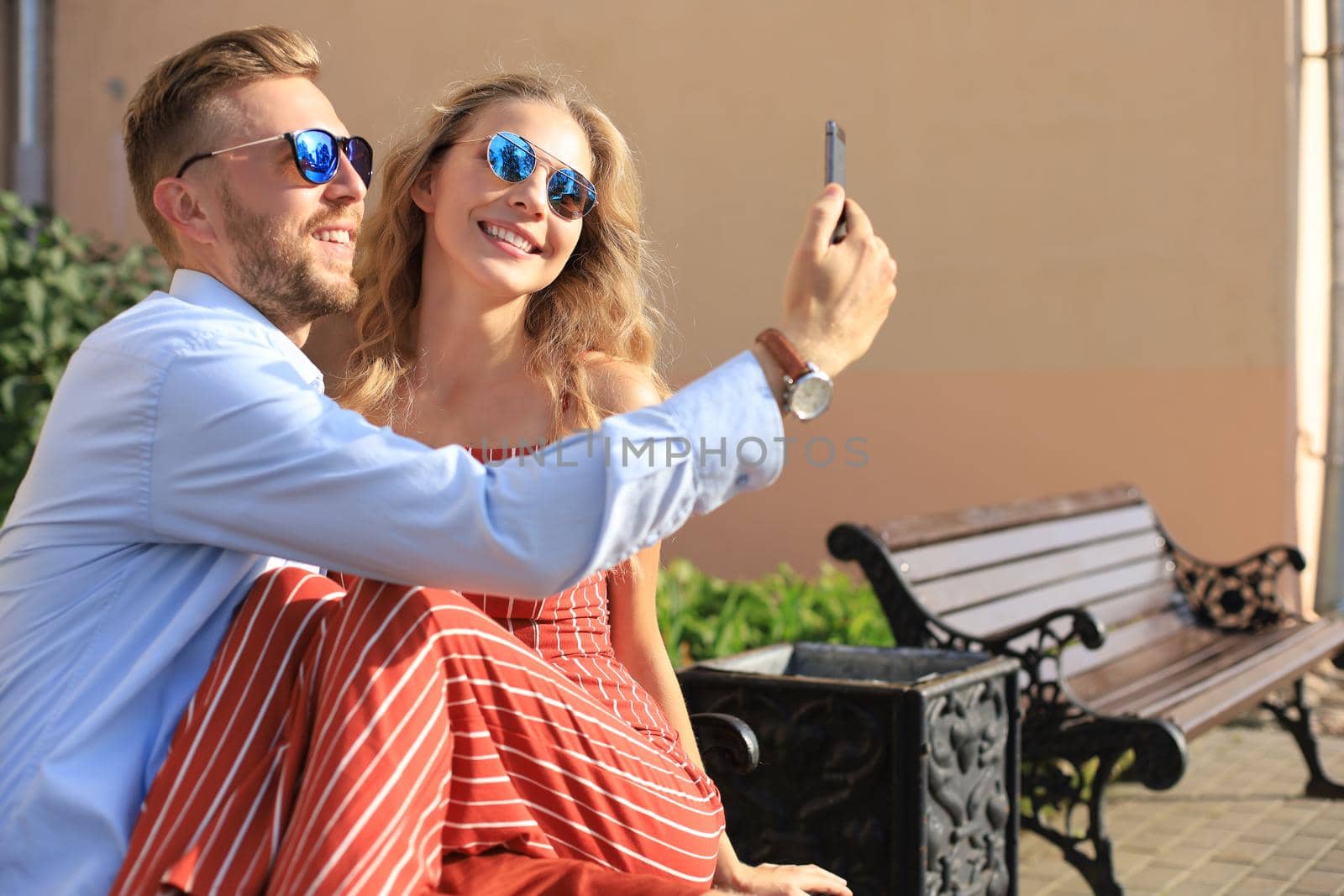 Romantic young couple in summer clothes smiling and taking selfie while sitting on bench in city street.