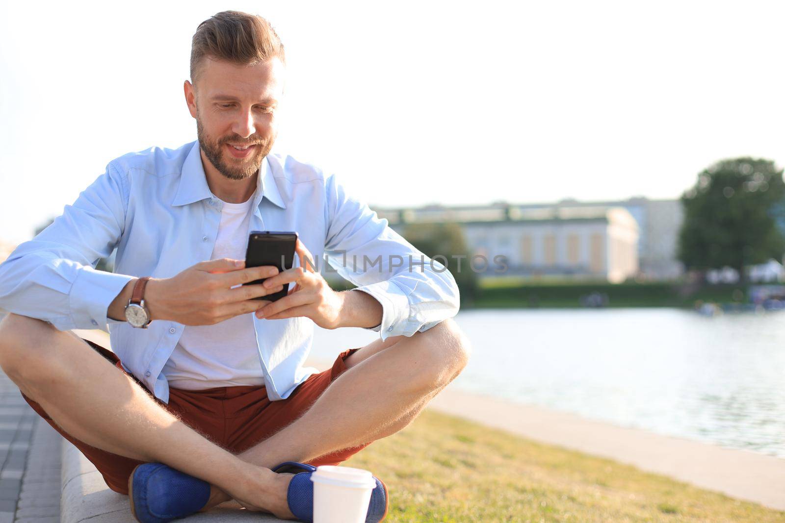 Happy young handsome man sitting on the bench outdoors and using smartphone.