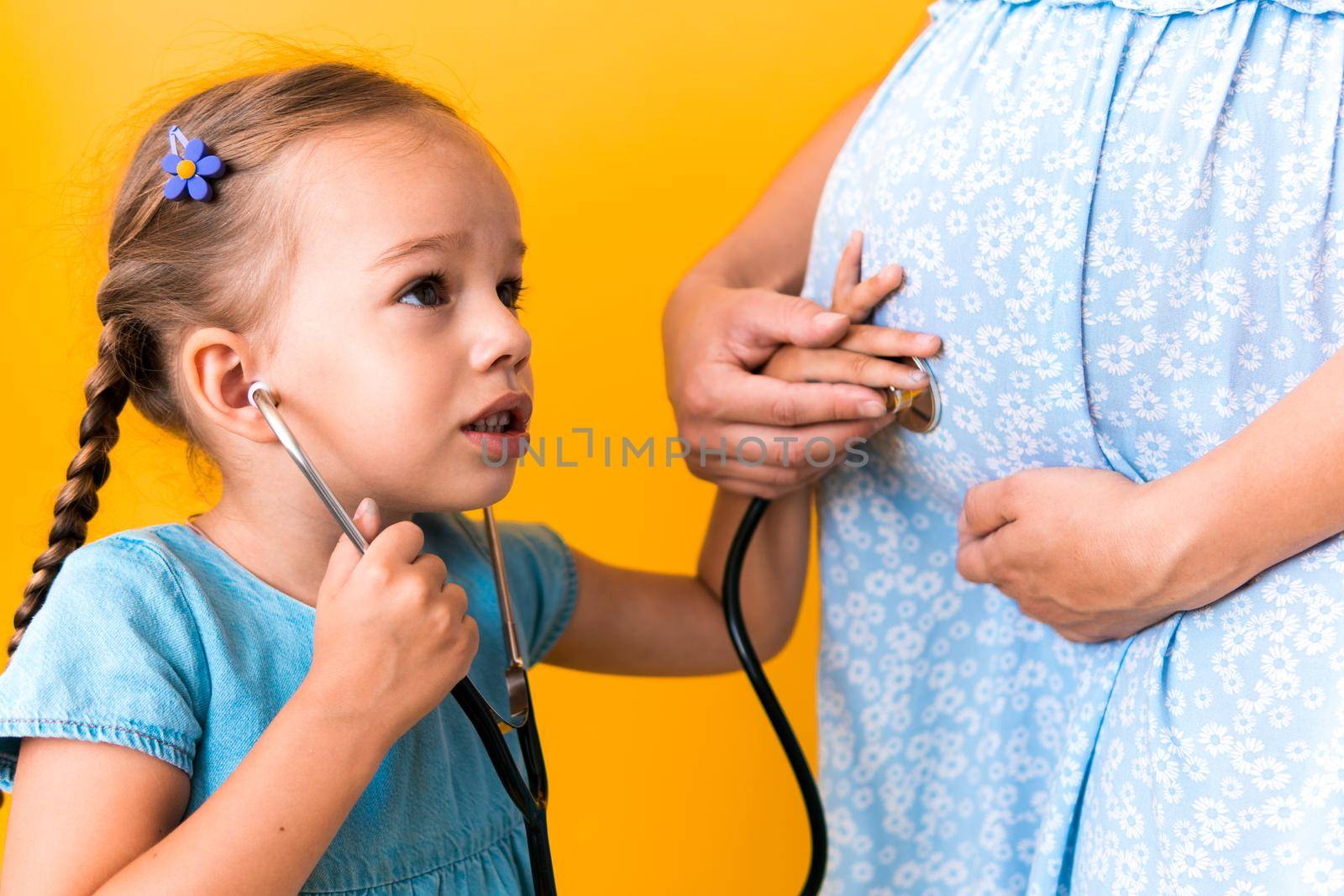 Motherhood, hot summer - croped portrait pregnant mother unrecognizable woman blue dress little Daughter girl sibling treat mom role play game stethoscope unborn big belly tummy on yellow background.