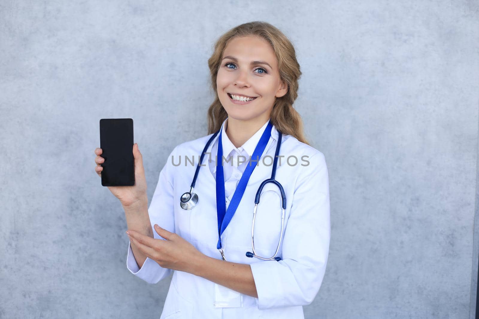 Female doctor smiling and showing a blank smartphone screen isolated over grey background. by tsyhun