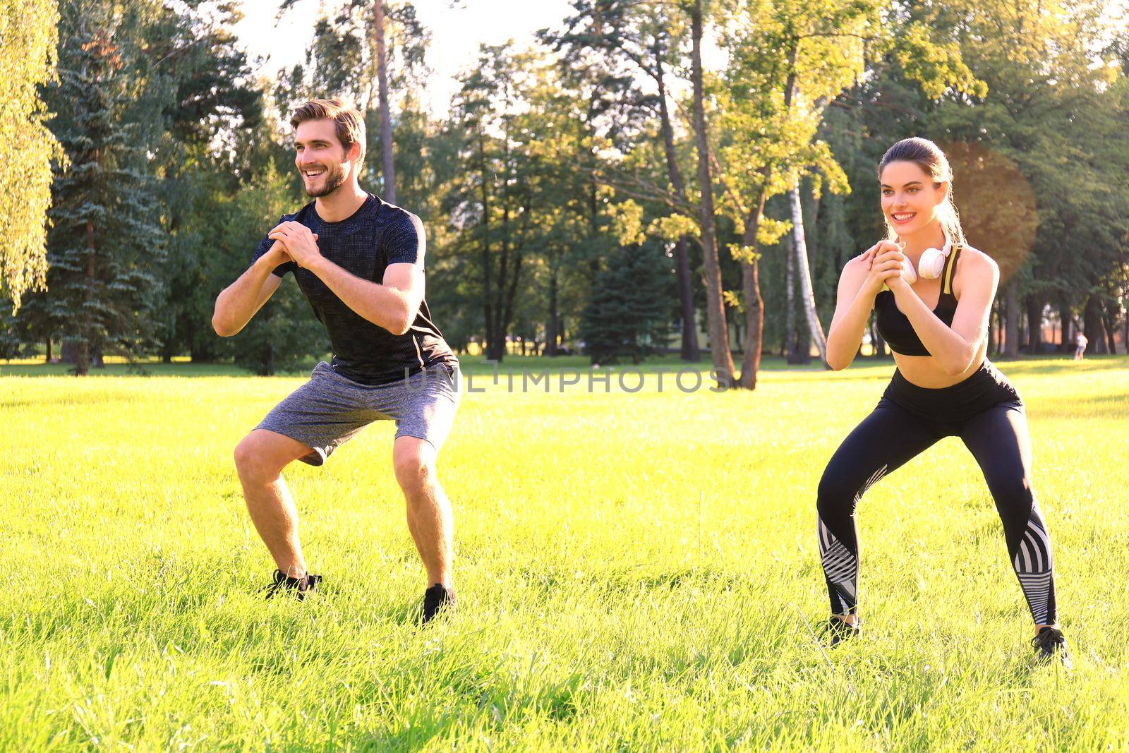 Young sporty man and woman doing workout and squatting together in green park during summer sunny day
