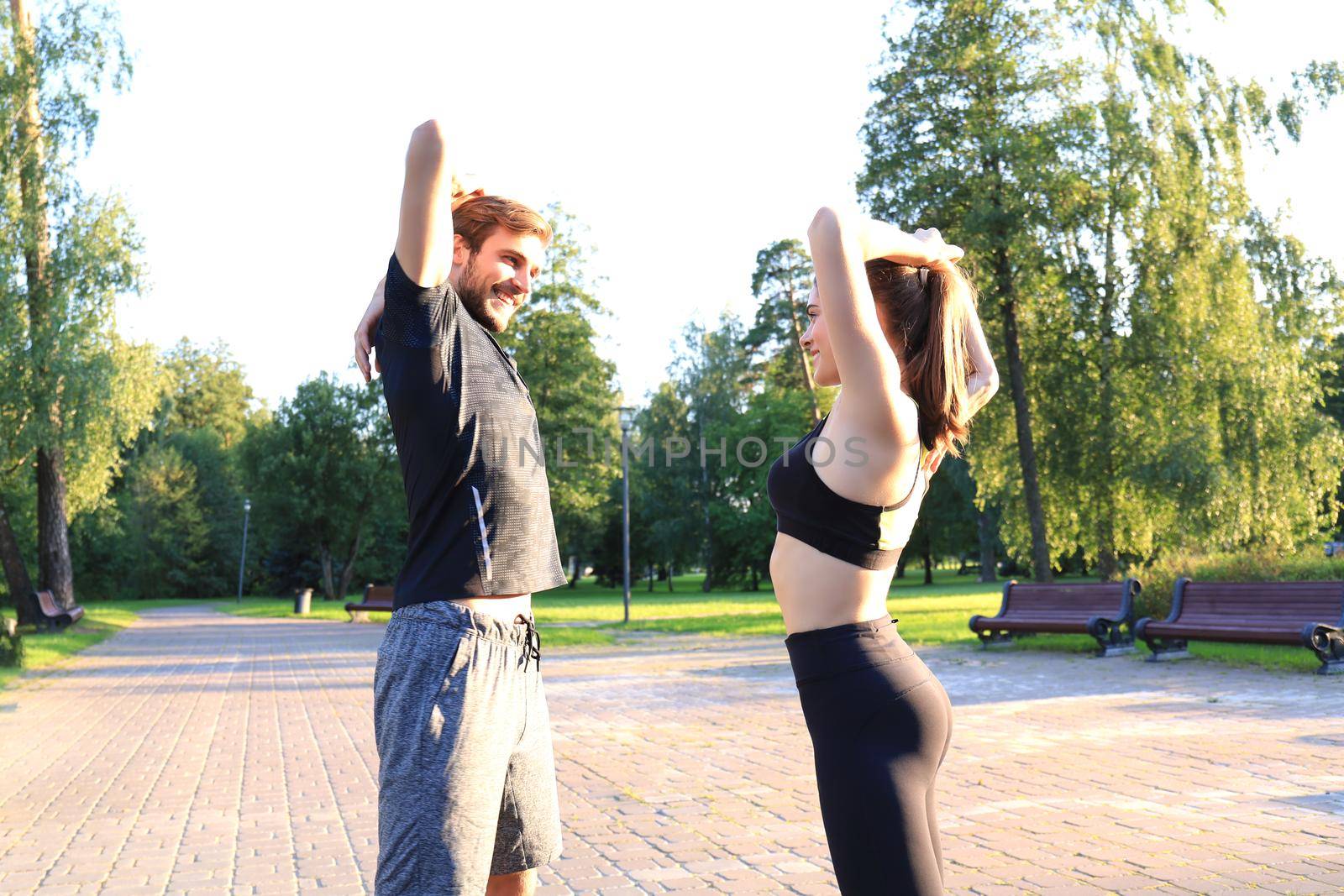Attractive couple doing stretch together and smiling while working out in park outdoors