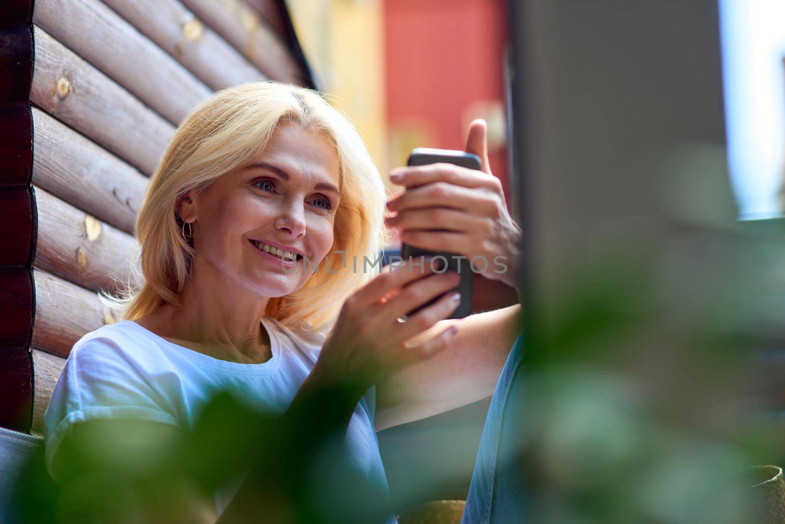 Pretty middle aged caucasian woman smiling to phone sitting on sunny terrace in apartment, selective focus. People spending time at home
