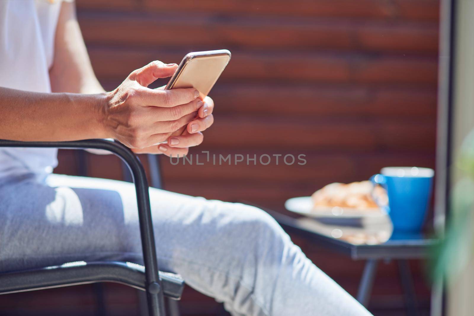 Woman holding smartphone in hands while resting on sunny terrace, cropped selective shot. People spending time at home
