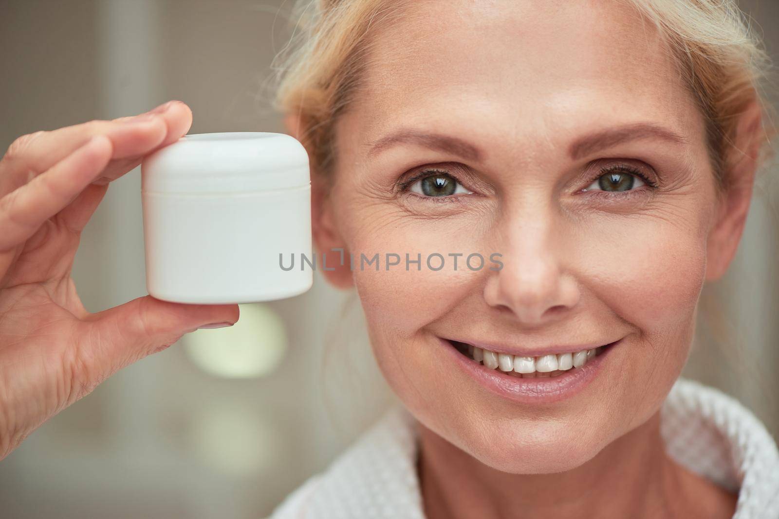 Portrait of beautiful middle aged caucasian woman with clean shiny skin holding plastic cosmetic cream bottle on near face while smiling at camera. Beauty, skincare and cosmetology concept