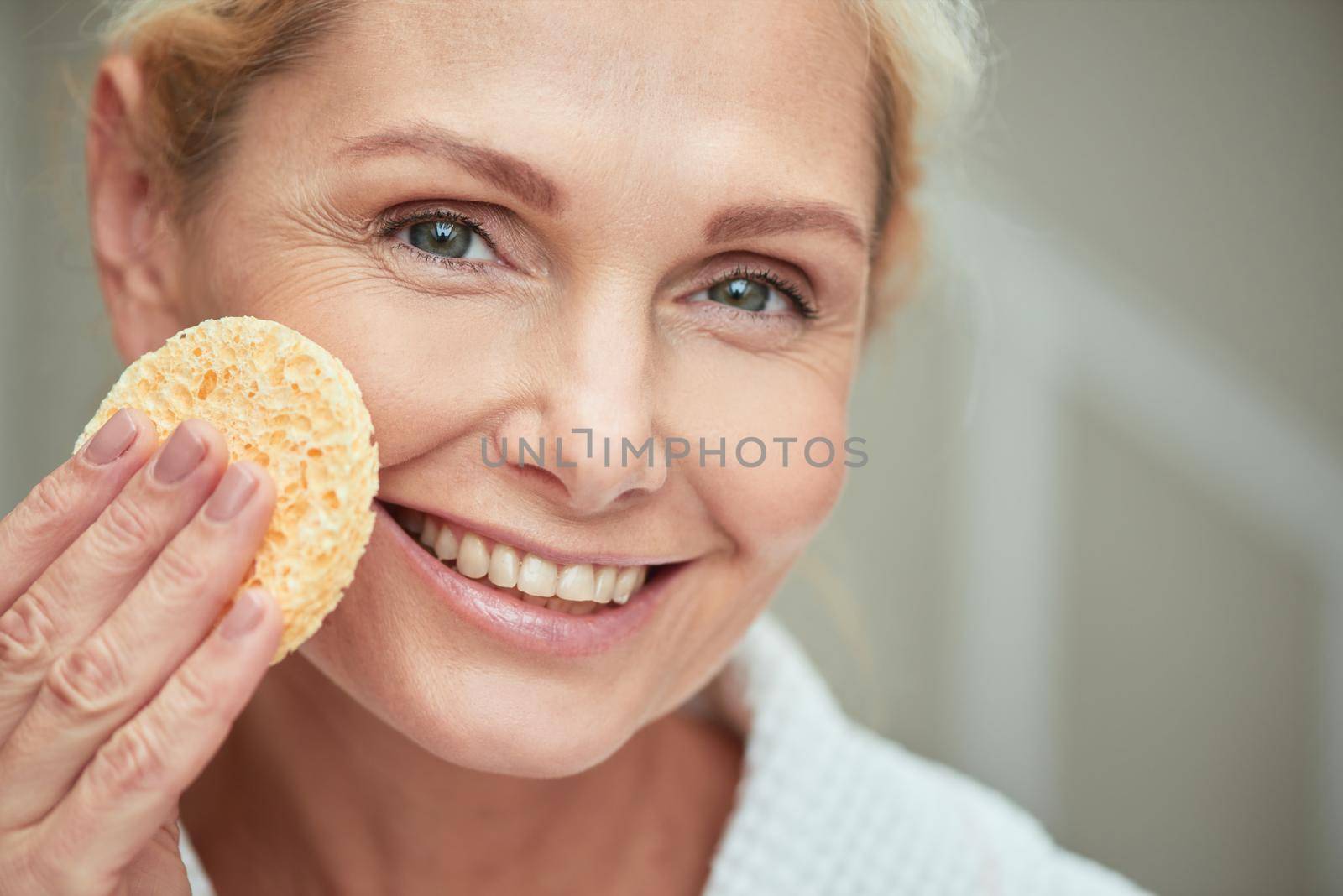 Smiling middle aged caucasian woman with clean shiny skin holding facial sponge while looking at camera. Beauty, skincare and cosmetology concept