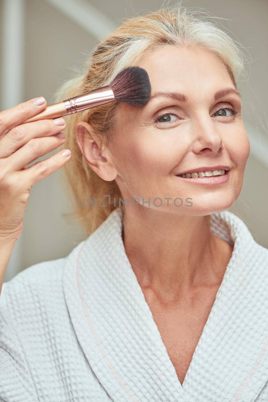 Smiling middle aged caucasian woman looking at camera and smiling while applying makeup brush on forehead. Beauty, skincare and cosmetology concept